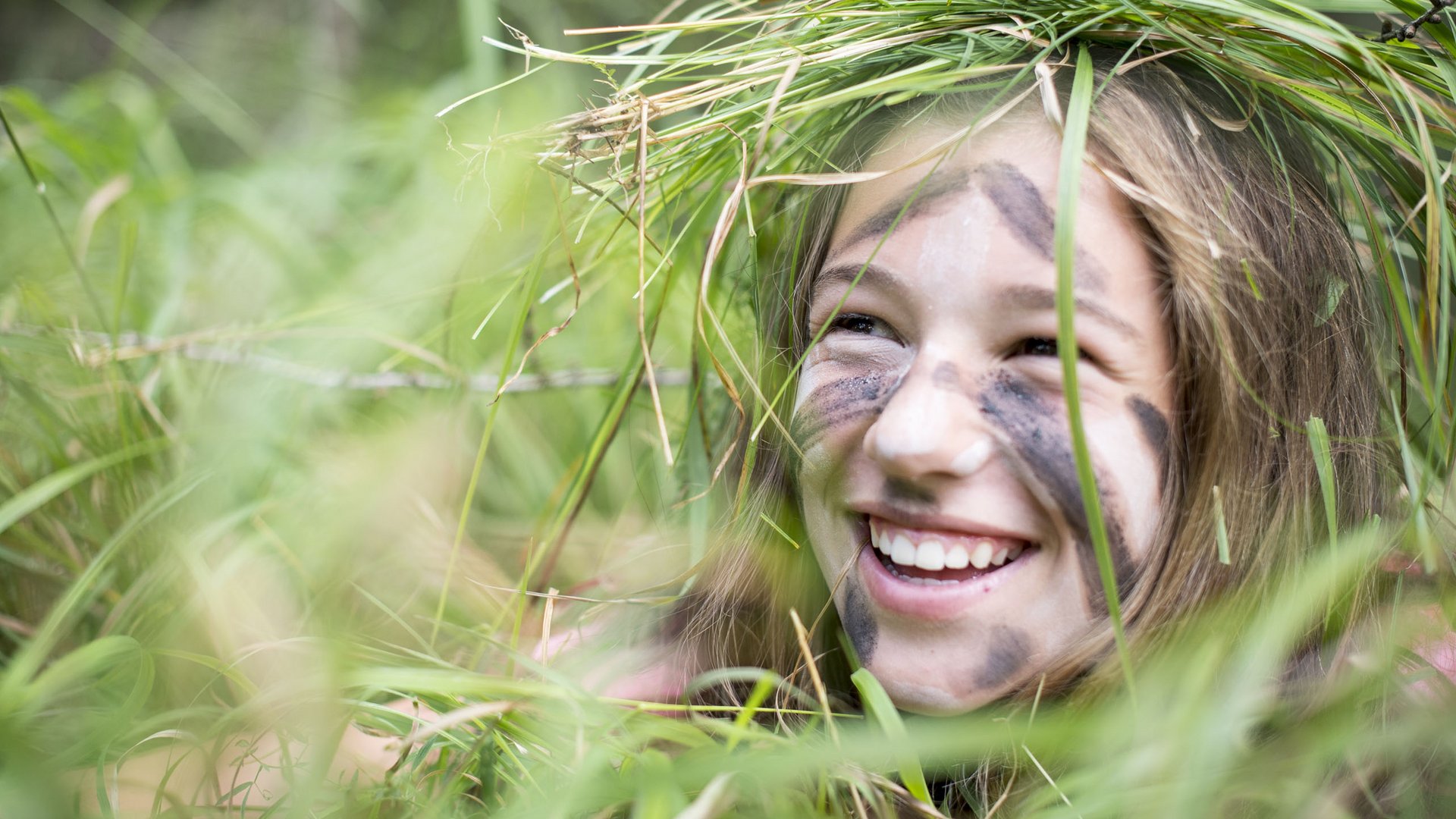 Kinderhotels in Südtirol 4-Sterne-Superior Lachendes Mädchen mit Tarnfarbe und Graskranz im Gesicht im Freien