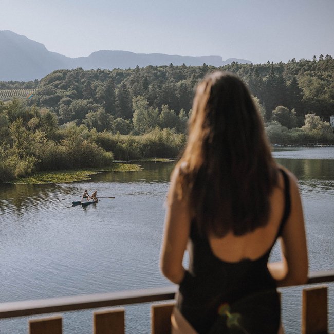 Hotels nahe dem Kalterer See 4-Sterne-Superior Frau schaut auf Fluss mit zwei Menschen in Ruderboot vor bewaldeten Hügeln
